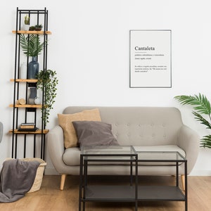 May include: A living room interior with a gray sofa, a gray armchair, a black metal shelf with wooden shelves, a glass coffee table, a potted palm tree, and a framed print with the word "Cantaleta" and its definition.