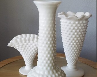 Puede incluir: Tres jarrones blancos con diseño hobnail de diferentes formas. El jarrón central es alto y cilíndrico, los otros tienen bordes acampanados o con volantes. Los jarrones están sobre una superficie de madera.