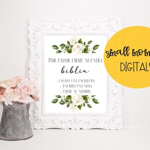May include: A white framed printable sign with a floral design and the text "POR FAVOR FIRME NUESTRA biblia LESCOJA UNA ESCRITURA ESCRIBA UNA NOTA FIRME SU NOMBRE" in black. The sign is sitting on a white surface with a small metal pitcher filled with pink flowers.