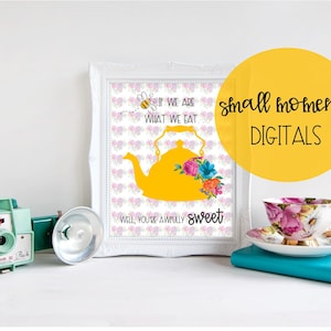 Puede incluir: Una impresión enmarcada en blanco con una silueta de tetera amarilla y detalles florales. La impresión tiene el texto "If we are what we eat, will you're awfully sweet." y una pequeña ilustración de abeja. La impresión está sobre una superficie blanca con una cámara vintage, una taza de té y un platillo, y un libro azul verdoso.