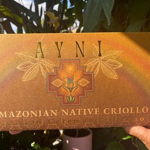 Puede incluir: Una caja rectangular con la palabra "AYNI" en letras doradas, con un diseño central con un arcoíris e ilustraciones botánicas. El texto "AMAZONIAN NATIVE CRIOLLO" está impreso debajo. La caja tiene una paleta de colores cálidos y terrosos.