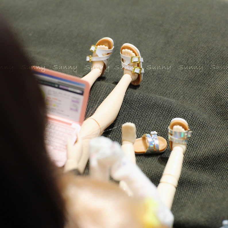 May include: Two dolls wearing sandals and sitting on a green fabric surface. One doll is wearing white sandals with gold buckles and the other doll is wearing silver sandals. The doll in the foreground is holding a laptop computer.
