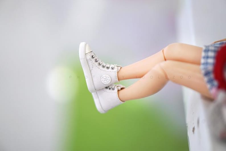 May include: A pair of white high-top sneakers with a white star logo on the side. The sneakers are worn by a doll with bare legs and a plaid skirt.