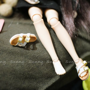 May include: A pair of white sandals with gold buckles on a doll's feet. The doll is wearing a black skirt and has long black hair.