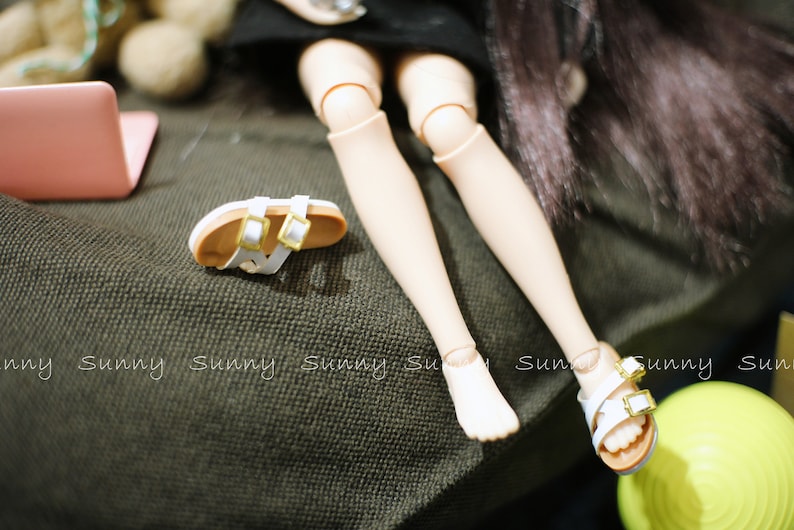 May include: A pair of white sandals with gold buckles on a doll's feet. The doll is wearing a black skirt and has long black hair.