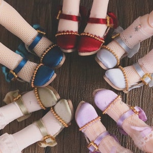 Può includere: Un set di otto scarpe in miniatura per bambole di diversi colori e stili. Le scarpe hanno fibbie dorate e sono decorate con perline. Le scarpe sono disposte in cerchio su una superficie di legno.