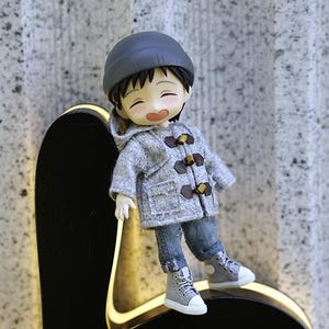 May include: A small gray doll wearing a gray hooded jacket with brown toggle buttons, blue jeans, and gray sneakers. The doll is smiling and has black hair. The doll is sitting on a black surface with a yellow outline.
