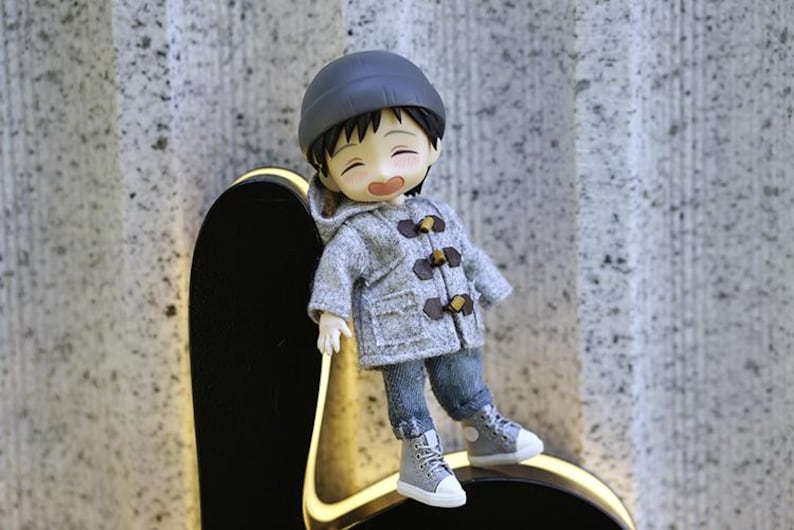 May include: A small gray doll wearing a gray hooded jacket with brown toggle buttons, blue jeans, and gray sneakers. The doll is smiling and has black hair. The doll is sitting on a black surface with a yellow outline.