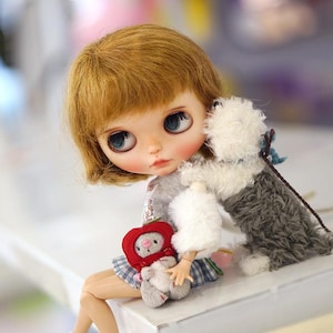 May include: A doll with blonde hair and blue eyes is sitting on a white surface. She is holding a red and white stuffed animal and a white fluffy dog is sitting next to her.
