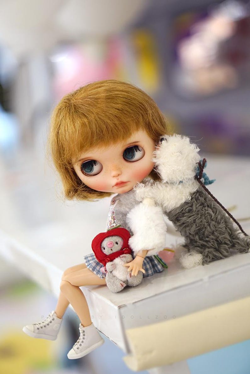 May include: A doll with blonde hair and blue eyes is sitting on a white surface. She is holding a red and white stuffed animal and a white fluffy dog is sitting next to her.