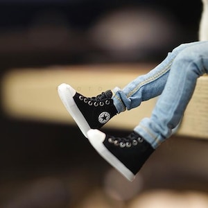 May include: A pair of black canvas high-top sneakers with white soles and a white star logo. The sneakers are worn by a person wearing blue jeans.