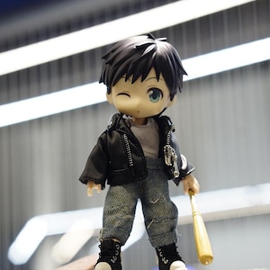 May include: A small, cartoon-style doll wearing a black leather jacket, blue jeans, and black sneakers. The doll is holding a wooden baseball bat and standing on a wooden skateboard.
