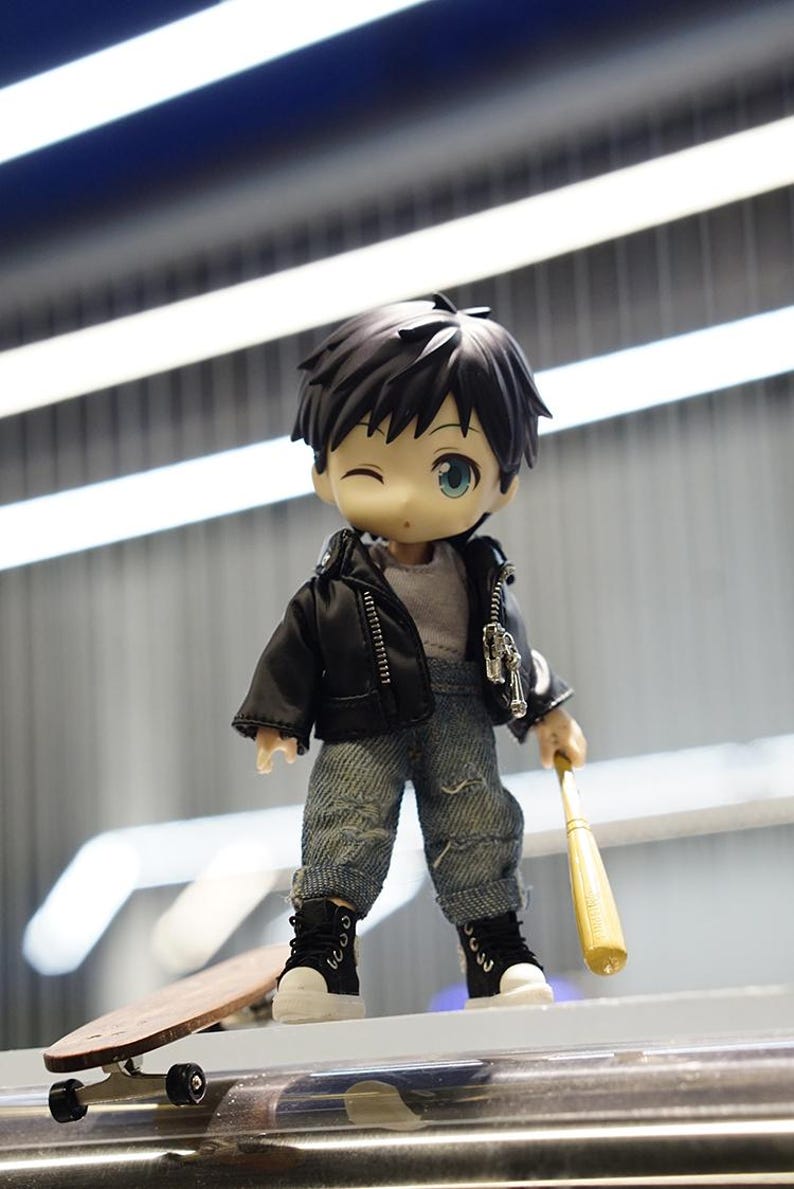 May include: A small, cartoon-style doll wearing a black leather jacket, blue jeans, and black sneakers. The doll is holding a wooden baseball bat and standing on a wooden skateboard.