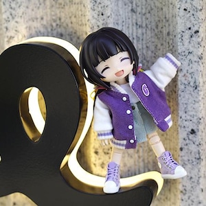 May include: A small purple and white doll with black hair and a big smile is standing on a black and gold sign. The doll is wearing a purple jacket with a white collar and white sleeves, a blue denim skirt, and purple shoes.