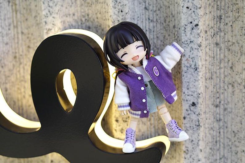 May include: A small purple and white doll with black hair and a big smile is standing on a black and gold sign. The doll is wearing a purple jacket with a white collar and white sleeves, a blue denim skirt, and purple shoes.