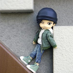 May include: A small doll wearing a green jacket, blue jeans, and a black beanie. The doll is wearing green sneakers and has black hair.