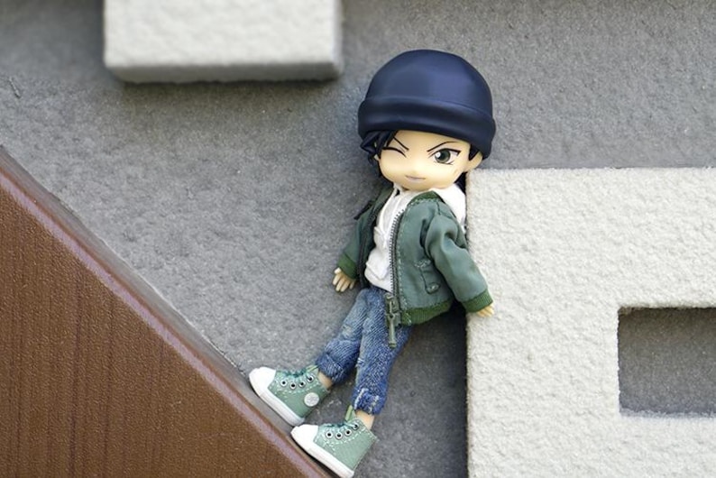 May include: A small doll wearing a green jacket, blue jeans, and a black beanie. The doll is wearing green sneakers and has black hair.