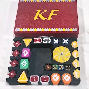 May include: A red storage box with the letters KF in yellow and a black game organizer tray containing various colorful tokens, including star-shaped pieces with numbers, yellow triangles, square markers, and four multi-colored dice.
