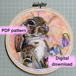 May include: A digital download PDF pattern for an embroidery project featuring a brown rabbit with a pink flower in its mouth and blue flowers in the background. The pattern is available for instant download.