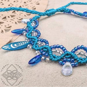 Könnte beinhalten: Eine türkisfarbene und blaue Makramee-Halskette mit silbernen Perlen und blauen Glasakzenten. Die Halskette hat ein gewebtes Design und ist mit kleinen, tropfenförmigen blauen Glasperlen verziert.