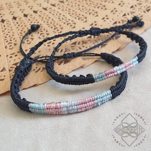 Könnte beinhalten: Zwei schwarze Freundschaftsarmbänder mit gewebten Streifen in Pink, Blau und Grau. Die Armbänder sind mit einem Knoten zusammengebunden.