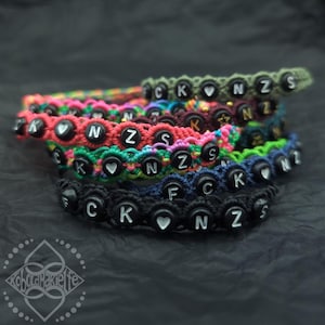 Könnte beinhalten: Eine Ansammlung farbenfroher Makramee-Armbänder in Pink, Grün, Blau und Schwarz. Jedes Armband hat schwarze Perlen mit weißen Buchstaben und Herzsymbolen. Die Buchstaben bilden verschiedene Wörter.