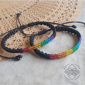 Könnte beinhalten: Zwei schwarze Freundschaftsarmbänder mit regenbogenfarbenen gewebten Streifen. Die Armbänder sind mit schwarzem Garn zusammengebunden.