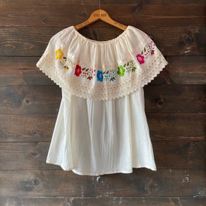 May include: A white cotton blouse with a ruffled neckline and short sleeves. The neckline is trimmed with white lace and features colorful embroidered flowers.