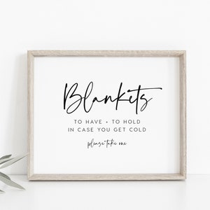 May include: A sign with the text "Blankets to have + to hold in case you get cold please take one" in a black cursive font on a white background. The sign is framed in a light wood frame.