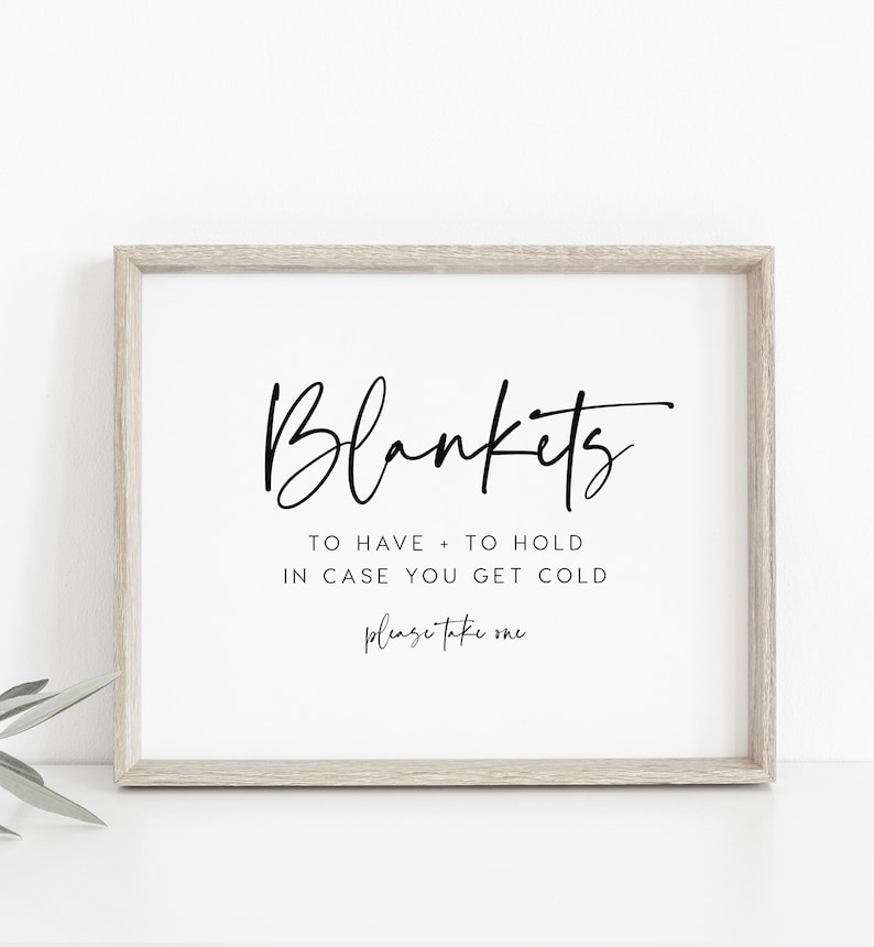 May include: A sign with the text "Blankets to have + to hold in case you get cold please take one" in a black cursive font on a white background. The sign is framed in a light wood frame.