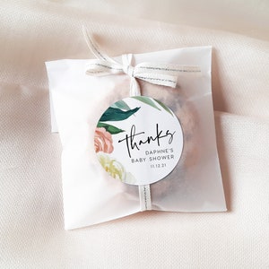 May include: A white favor bag with a round, floral thank you sticker. The sticker says "Thanks" in cursive, "Daphne's Baby Shower" and "11.12.21".