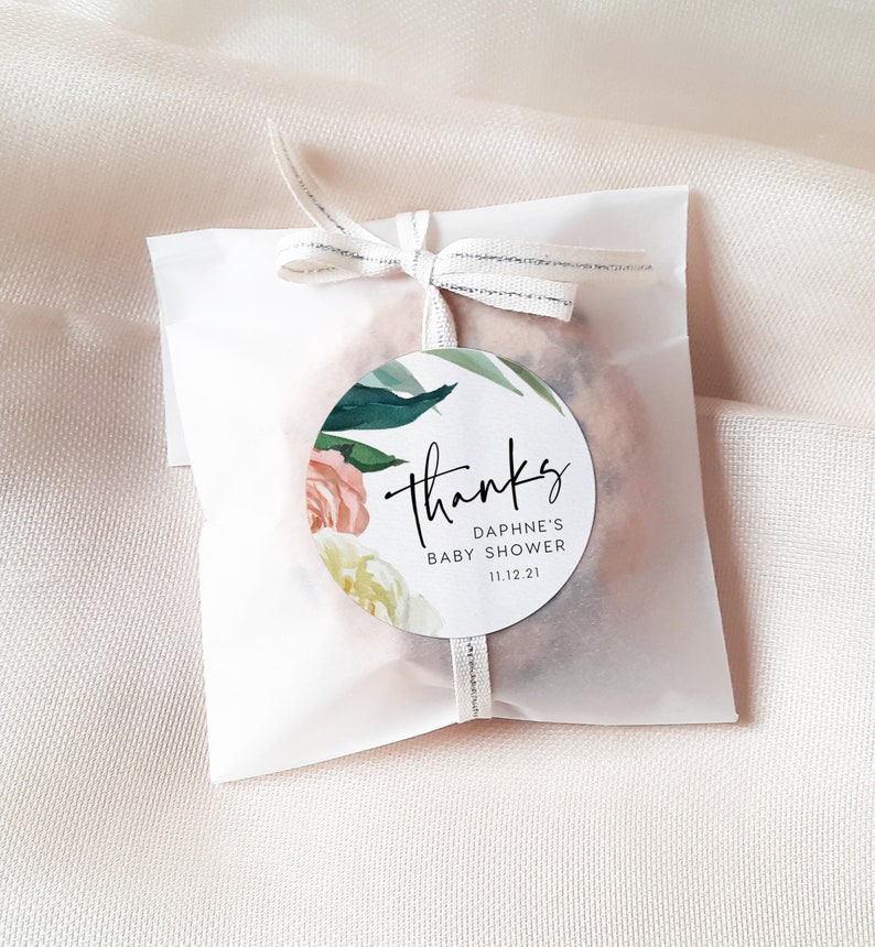 May include: A white favor bag with a round, floral thank you sticker. The sticker says "Thanks" in cursive, "Daphne's Baby Shower" and "11.12.21".