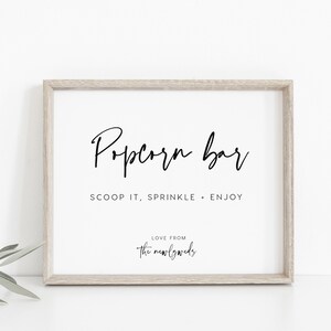 May include: A framed sign with the text "Popcorn Bar" in black cursive font. Below the text, it reads "Scoop it, Sprinkle + Enjoy". At the bottom of the sign, it says "Love from the newlyweds".