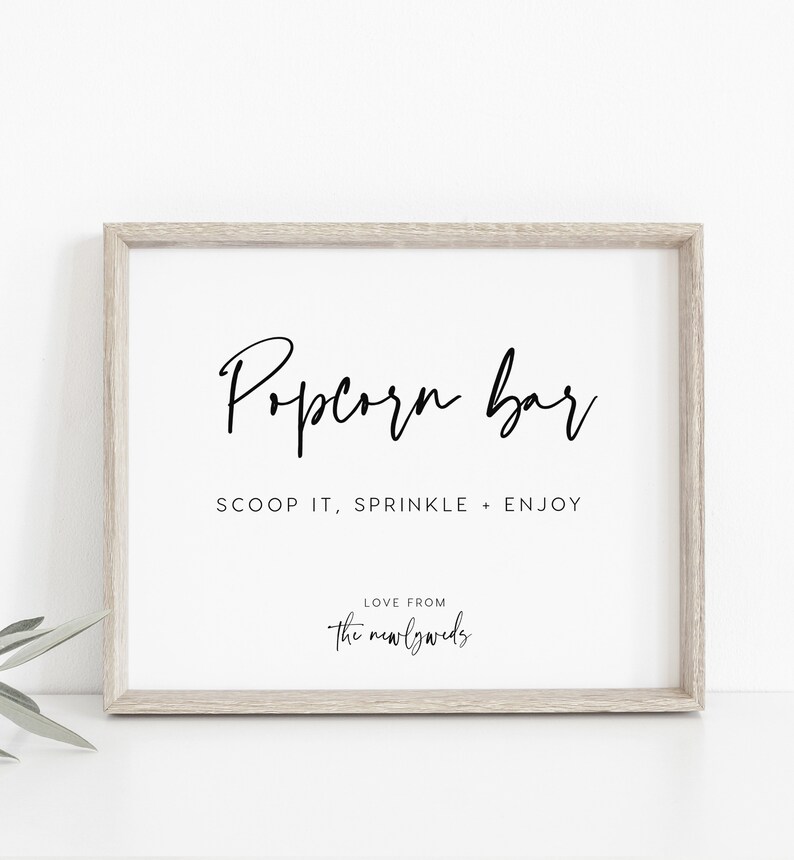May include: A framed sign with the text "Popcorn Bar" in black cursive font. Below the text, it reads "Scoop it, Sprinkle + Enjoy". At the bottom of the sign, it says "Love from the newlyweds".