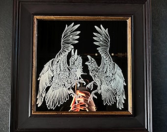 May include: Square framed mirror with a dark brown wooden frame. The mirror features a detailed etched design of two birds facing each other, with their wings spread. The mirror's background is black, and the frame has a gold inner edge.