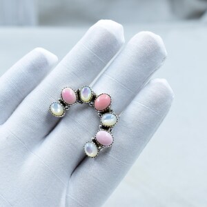 May include: A silver ring with a curved design, set with alternating pink and opalescent oval gemstones. The ring is displayed on a white surface.