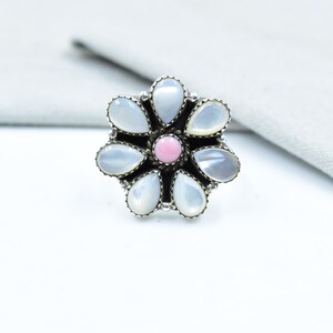 May include: A silver ring with a floral design. The ring has pear-shaped, pale stones encircling a pink central stone. The ring is set against a white backdrop with grey fabric in the background.