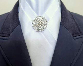 May include: A white satin stock tie worn with a dark navy blue blazer. The stock tie is secured at the center with a round, silver-toned brooch featuring a floral design encrusted with clear, sparkling crystals that catch the light.