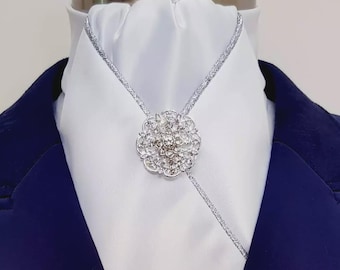 May include: A silver-toned bolo tie featuring a circular, floral-shaped centerpiece encrusted with clear, sparkling crystals. The tie consists of a thin, textured silver cord that loops through the central ornament, resting against a white ascot.