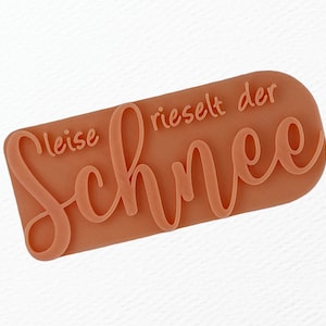 May include: A light brown rubber stamp with the text "leise rieselt der Schnee" in a cursive font.