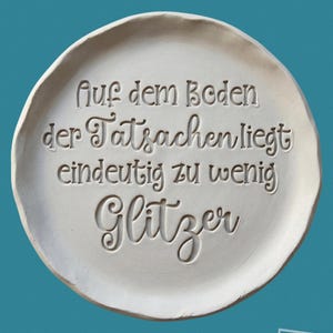 Könnte beinhalten: Eine runde, cremefarbene Keramikplatte mit dem geprägten deutschen Text "Auf dem Boden der Tatsachen liegt eindeutig zu wenig Glitzer". Die Platte hat einen leicht unregelmäßigen Rand, vor türkisfarbenem Hintergrund.