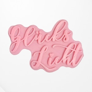 Könnte beinhalten: Ein rosa Stempel mit den Worten "Glück's Licht" in einer kursivschrift.