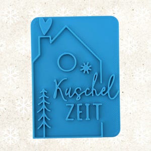 Könnte beinhalten: Ein blauer rechteckiger Stempel mit einem Haus-, Herz- und Baumdesign. Der Stempel enthält die Wörter "Kuschel ZEIT" in einer Schreibschrift. Das Design hat ein Winter- oder Feiertags-Thema.
