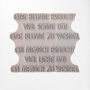 Op de afbeelding: Een wit 3D-geprint bord met de tekst "eine BLUME BRAUCHT Viel sonne um eine BLUME ZU WERDEN ein Mensch BRAUCHT VIEL LIEBE UM ein Mensch ZU WERDEN."