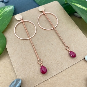 May include: A pair of rose gold-toned earrings featuring a circular hoop attached to a vertical wire-wrapped bar. Each earring terminates in a small, faceted, teardrop-shaped magenta gemstone suspended from the base of the metal bar.