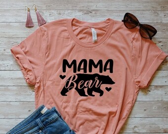 May include: A peach-colored t-shirt with the text "Mama Bear" in black lettering and a black bear silhouette. The shirt is folded and laid on a wooden surface with a pair of blue jeans, a leopard print clutch, and a pair of sunglasses.