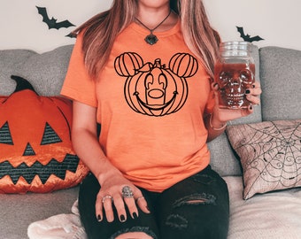 Puede incluir: Camiseta naranja con un contorno negro de una calabaza con orejas de Mickey Mouse. La camiseta se usa con vaqueros negros. También se ven una almohada decorativa de calabaza y un vaso con forma de calavera, lo que sugiere un tema de Halloween.