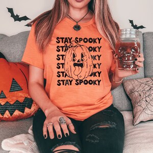 May include: A woman wearing an orange t-shirt with the text "Stay Spooky" repeated multiple times. The shirt features a black and white illustration of a smiling pumpkin with a tongue sticking out. The woman is holding a glass jar with a skull design.