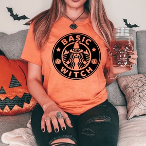May include: A woman wearing an orange t-shirt with a black graphic of a witch and the text "Basic Witch" in a coffee cup logo. She is holding a glass with a skull design.