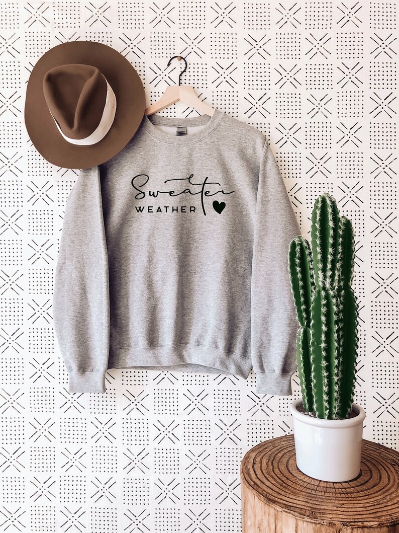 May include: Gray sweatshirt with black text that reads "Sweater Weather" with a black heart. The sweatshirt is hanging on a wooden hanger against a white background with a black geometric pattern.